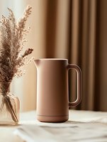 Minimalist beige ceramic pitcher on white tablecloth with dried pampas grass bouquet in glass vase against neutral brown background, modern home decor styling.