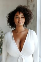Young African American woman with natural curly hair wearing white wrap dress against neutral background. Soft natural lighting creates elegant portrait.