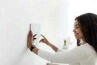 Young Hispanic woman adjusting smart home thermostat on white wall, wearing white sweater, modern energy-efficient technology for comfortable living.