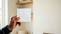 Close-up of a hand holding a blank white card or paper against a neutral beige wall background, suitable for mockups, advertising, and promotional designs.