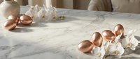 Elegant copper eggs scattered on marble surface with white orchid flowers in soft natural lighting for luxury Easter decoration and spring celebration.