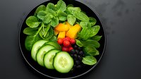 Fresh salad ingredients arranged on black plate with sliced cucumber, kumquats, strawberries, blackberries and green spinach leaves, viewed from above on dark background.