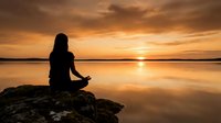 Woman meditating on rock at sunset beach, silhouetted against golden sky and calm water reflection, mindfulness and relaxation concept.