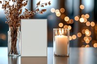 White blank frame mockup with glowing candle in glass holder and dried flowers in vase against blurred bokeh lights background. Cozy home decor setting.