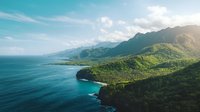 Dramatic coastal landscape with lush green mountains meeting turquoise ocean waters under soft sunlight and scattered clouds, showcasing pristine wilderness coastline.