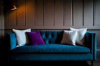 Modern teal velvet sofa with purple and white throw pillows against dark paneled wall, illuminated by dramatic directional lighting creating moody atmosphere.