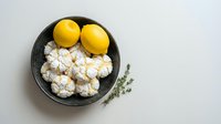 Fresh lemons and cauliflower florets arranged in dark bowl with thyme herbs on white background for healthy cooking and meal preparation concepts.