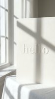 White embossed greeting card with hello text standing near sunlit window, casting soft shadows on linen surface, minimal elegant style.