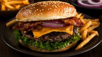 Juicy cheeseburger with crispy bacon, melted cheddar, fresh lettuce, and red onion rings served on sesame bun with golden french fries on black plate.