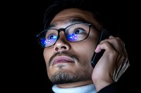 Asian male professional wearing glasses with blue light reflection while talking on smartphone, dramatic low key lighting creates modern tech atmosphere.