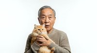 Senior Asian man with gray hair holding an orange tabby cat against his chest, wearing a beige knit sweater, isolated on white background for pet care and lifestyle content.