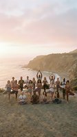 Large group of diverse men and women practicing yoga together on sandy beach at sunset with dramatic coastal cliffs in background. Wellness and fitness retreat.
