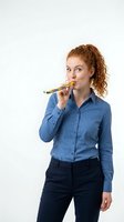 Young woman with curly reddish-brown hair and fair complexion wearing blue button-up shirt and dark trousers, chewing on colorful pencils against white background for creative concepts.