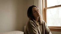 Young Asian woman in cozy beige sweater sitting peacefully by window with natural sunlight streaming in, expressing contentment and relaxation at home.