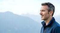 Middle-aged Caucasian male in blue jacket smiling outdoors with misty mountain landscape background, suitable for lifestyle, wellness, and corporate marketing materials.