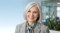Mature woman with white hair, fair complexion, wearing gray blazer and colorful scarf in modern office setting. Professional portrait for business, corporate, and leadership concepts.