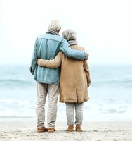 Senior couple embracing on beach looking at ocean waves, elderly man and woman in casual clothing enjoying romantic seaside moment together in golden light.