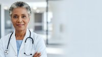 Confident mature African American female doctor with stethoscope and crossed arms smiling in modern hospital corridor for healthcare marketing.