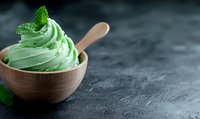 Creamy mint ice cream swirl in wooden bowl with fresh mint leaf garnish on dark stone background, soft-serve dessert with natural green color.