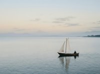 Lone sailor on a small wooden sailboat gliding across calm water at dusk, soft pastel sky and distant mountains reflecting serenity.