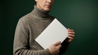 Middle-aged Caucasian man in brown turtleneck sweater holding blank white paper document against dark green background for business presentations.