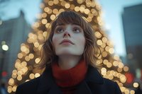 Young woman in red turtleneck and black coat gazing upward against blurred Christmas lights background creating magical bokeh effect in evening urban setting.