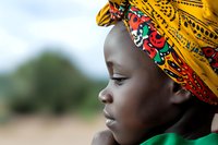 Profile of young African girl wearing vibrant yellow and floral traditional headwrap, looking thoughtfully into distance against blurred natural background.