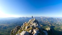 Lone hiker stands triumphantly on rocky mountain peak overlooking vast mountain range and valley landscape against clear blue sky on sunny day. Adventure travel concept.