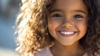 Young girl with curly brown hair and bright genuine smile showing white teeth against soft blurred background, radiating joy and innocence in natural lighting.