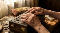Elderly hands gently touching vintage wooden jewelry box with brass hardware, surrounded by old photographs and warm sunlight streaming through window.