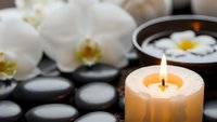 Spa wellness scene with burning candle, white orchid flowers, massage stones and floating frangipani in water bowl creating peaceful relaxation atmosphere.
