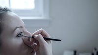 Young woman applying black eyeliner makeup with precision brush near bright window in modern bathroom for daily beauty routine and cosmetic application.