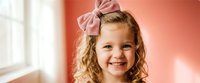 Young blonde girl with curly hair wearing pink bow headband smiling cheerfully in bright indoor setting with warm coral background lighting.