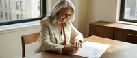 Mature blonde businesswoman in beige suit signing contract documents at wooden desk in bright modern office with city view windows.
