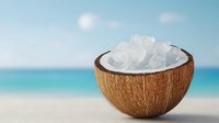 Coconut shell bowl filled with white sea salt crystals on tropical beach with turquoise ocean and blue sky background for spa wellness cooking concepts.