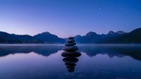 Zen stone stack balanced on calm lake water at twilight with mountain silhouettes and purple sky reflection creating peaceful meditation atmosphere.