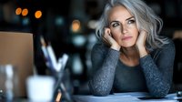 Mature woman with gray hair and fair complexion, wearing a dark gray sweater, resting her face on her hands at a desk with a computer and coffee cup, contemplative expression in an office setting.