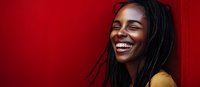 Young African American woman with dreadlocks wearing yellow top laughing joyfully against vibrant red wall background, expressing genuine happiness and positive emotions.