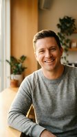 Happy middle-aged Caucasian man in grey sweater smiling at camera in cozy home interior with plants and warm natural lighting for lifestyle content.