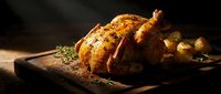 Golden roasted whole chicken with herbs and potatoes on wooden cutting board, warm lighting creates cozy kitchen atmosphere for culinary content.