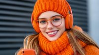 Young Caucasian woman in bright orange knit beanie and matching scarf smiling while wearing headphones and glasses. Modern winter fashion lifestyle portrait.