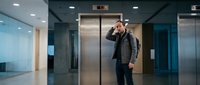 Stressed businessman talking on phone in modern office lobby near elevators during busy workday.