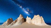 Dramatic limestone rock formations under starry night sky with Milky Way galaxy stretching across deep blue space. Desert landscape meets ocean horizon.