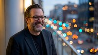 Smiling middle-aged Caucasian businessman with beard and glasses in dark suit jacket against blurred city lights background at evening office window.