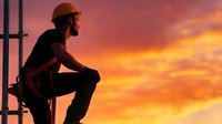 Silhouette of construction worker in safety helmet sitting on height against dramatic orange sunset sky, industrial concept for workplace safety.