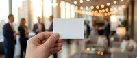 Hand holding blank white business card in modern office lobby with blurred professionals and warm lighting for corporate branding mockup.