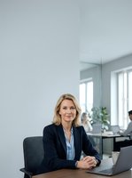 Mature woman professional in dark blazer and light blue shirt sitting at desk with laptop in modern office space with white walls and natural light from windows.