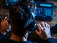 Caucasian male gamer wearing headphones in dark room with blue monitor glow, rear view. Dramatic lighting creates moody atmosphere for gaming and esports concept.