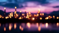 Blurred city lights create magical bokeh effect reflecting in calm water at twilight, with purple and orange sunset sky creating dreamy urban landscape.