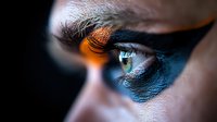 Dramatic close-up of green eye with vibrant blue eyeshadow and orange light reflection, showcasing intense makeup artistry and beauty photography techniques.